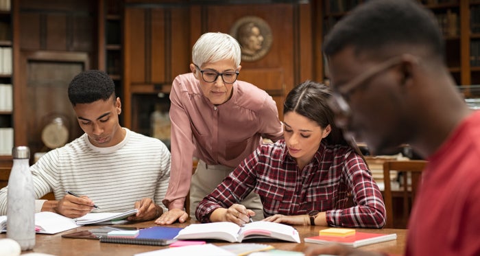 Senior woman teacher working with college students in library. Graduates consulting with their lecturer informally during a break in library. Group of students studying with their professor.