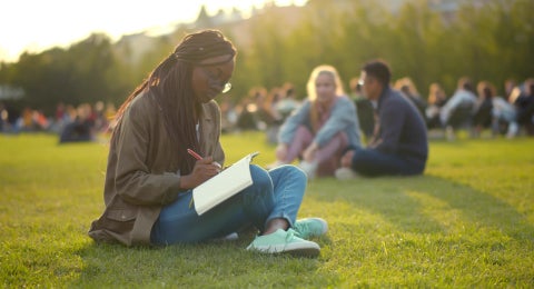 Pensive african female student writing in exercise book and doing homework in summer park. Portrait of beautiful afro-american college woman preparing for exam relaxing on campus lawn.