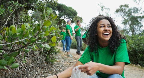 A student participating in a trash clean up smiling as she gets a plastic bottle out of a forested area.