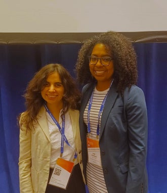 Two women stand side-by-side at a conference wearing name badges, smiling at the camera.