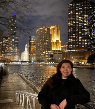 Carlia Lopez standing on the shore of the Chicago river with the city skyline in the background.