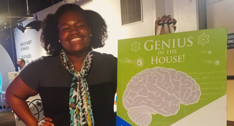Woman smiles at camera next to a poster that reads, "Genius in the House."