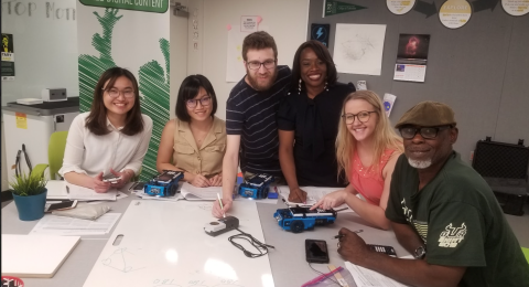 Six people gathered around a table in a classroom using calculators and collaborating on a whiteboard. Everyone is looking up at the camera and smiling.