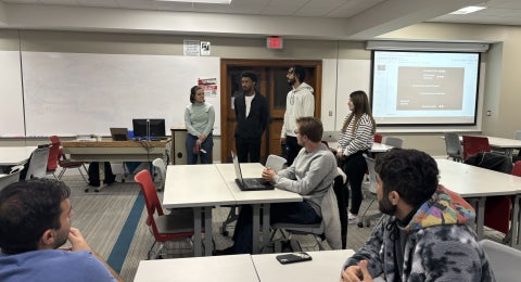 Students participating in a STEM CONNECT activity in a classroom with three students seated at desks looking at four students standing and speaking at the front of the room.