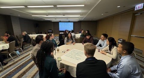 Students sitting at a round table in a meeting room participating in a group activity at a professional meeting.